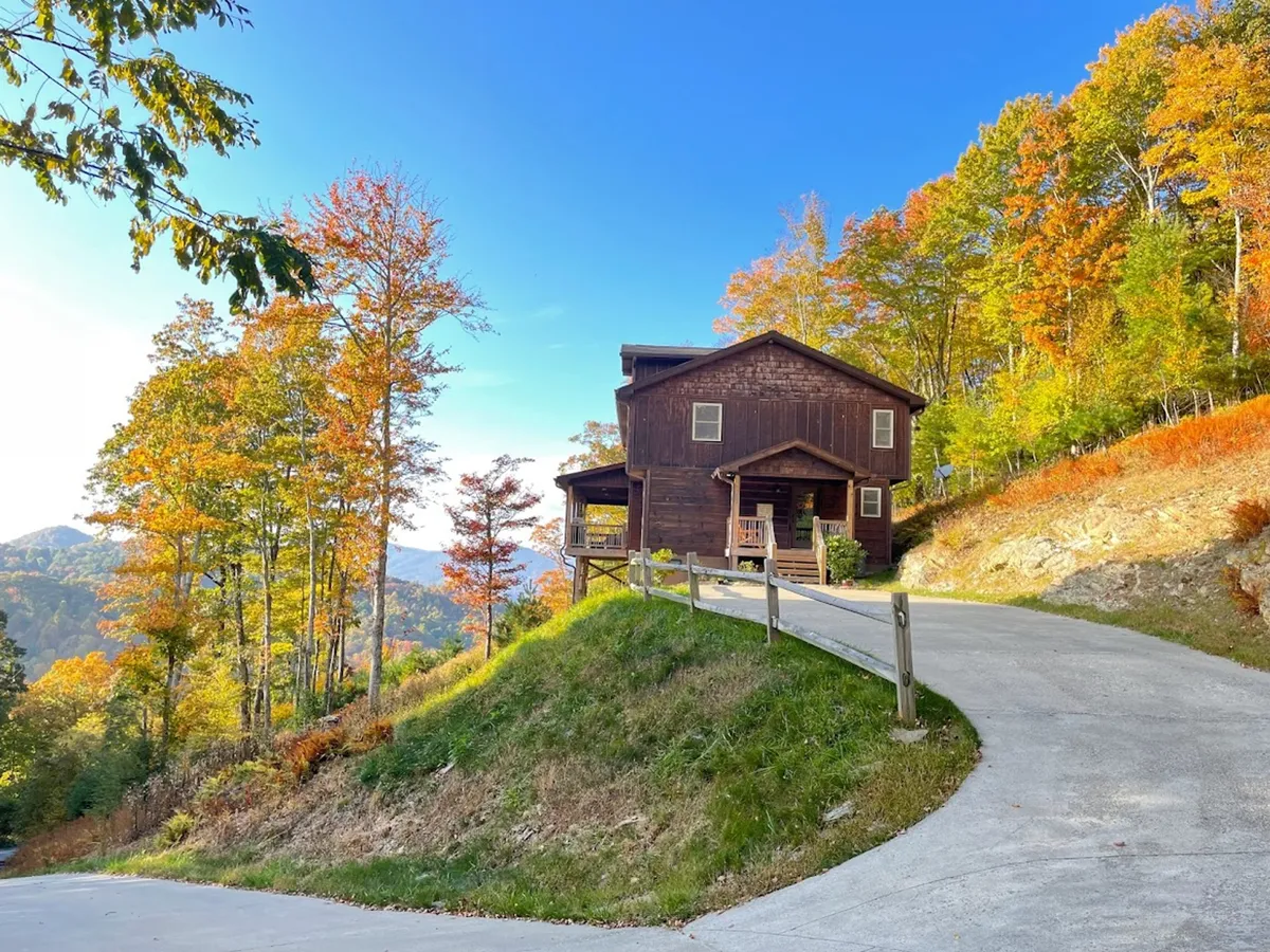 A charming wooden cabin nestled among colorful trees, with a winding driveway leading up to the front. The wraparound porch offers a perfect vantage point for admiring mountain sunsets, while the surrounding landscape hints at endless nature trails waiting to be explored.