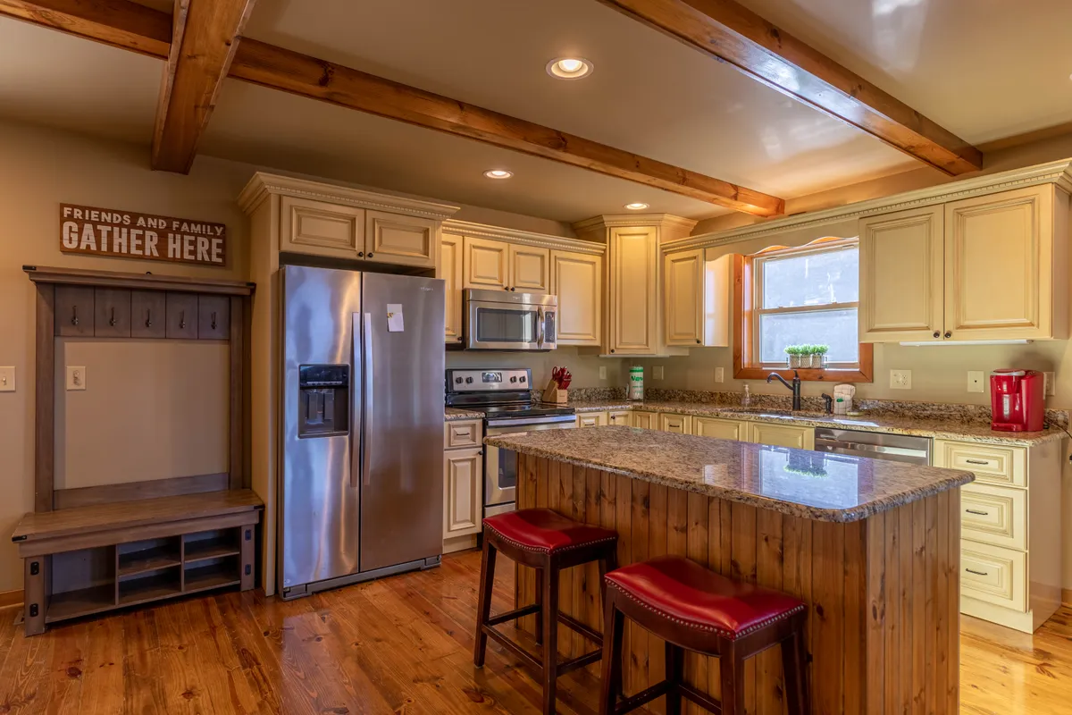 This image showcases a warm, inviting kitchen space complete with granite countertops, modern stainless steel appliances, wooden beams, and red bar stools at a kitchen island. The layout reveals both preparation and gathering areas, emphasizing the kitchen's dual purpose for cooking and socializing.