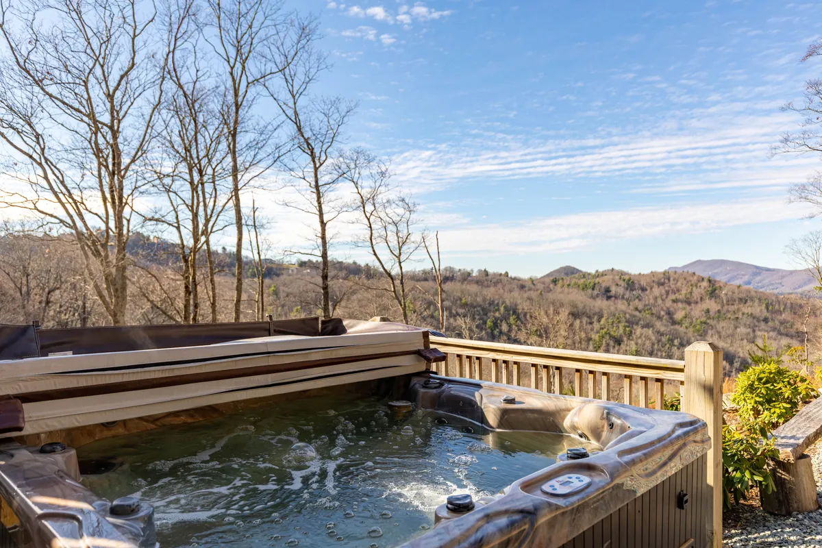This exterior image shows a hot tub set against the backdrop of bare trees and rolling mountains, offering both seclusion and a panoramic view. The tranquil mood is enhanced by the opportunity to soak, stargaze, or watch the sun rise and set from the warmth of the tub.