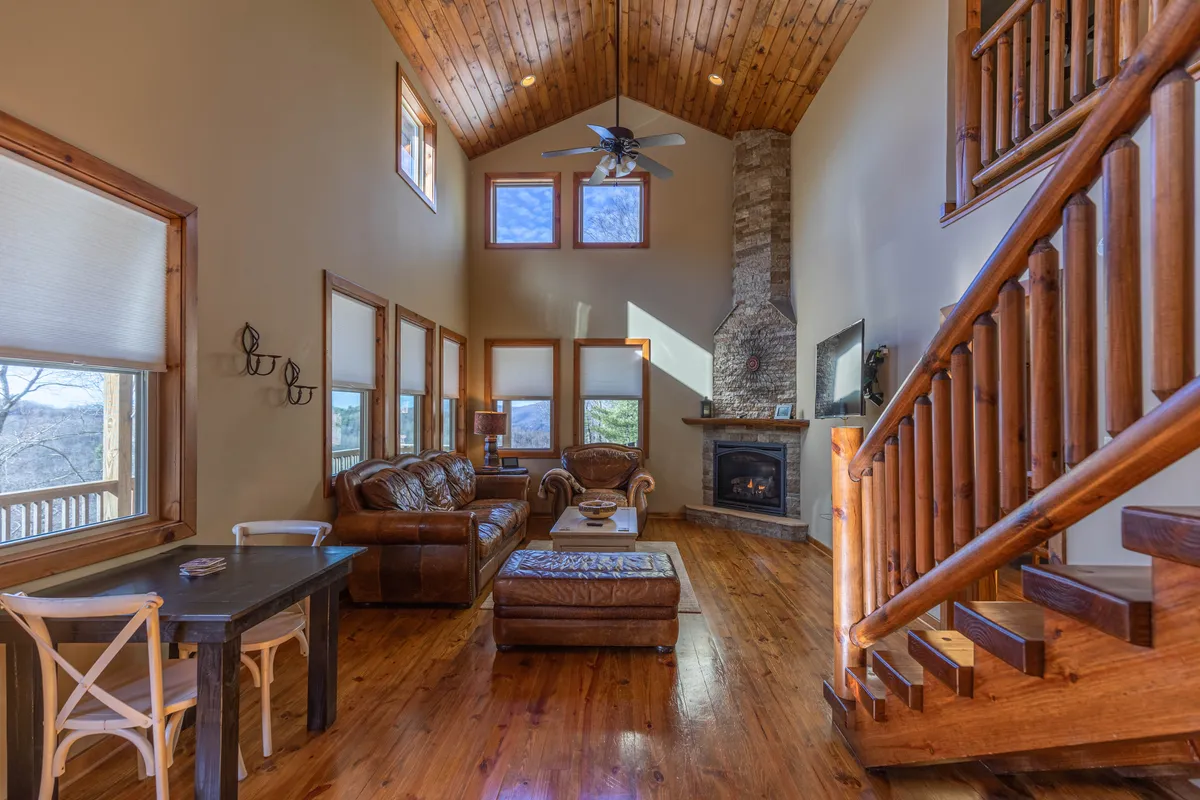 A spacious great room featuring a soaring vaulted wooden ceiling, a massive stone fireplace at the center, comfortable leather sofas arranged for conversation or relaxation, and a dining table in the background. Natural light streams in from large windows, enhancing the warm ambiance created by the log-and-beam accents and hardwood floors.