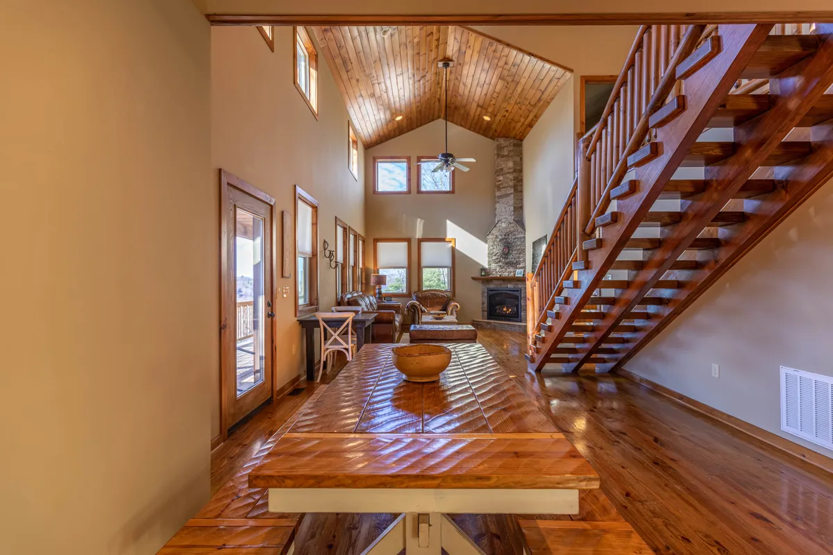 A generous dining area featuring a large wooden table that seats a crowd, an adjacent staircase leading to the loft, a cozy fireplace, and expansive windows offering sunlight and scenic views. The setting is primed for group meals and socializing.