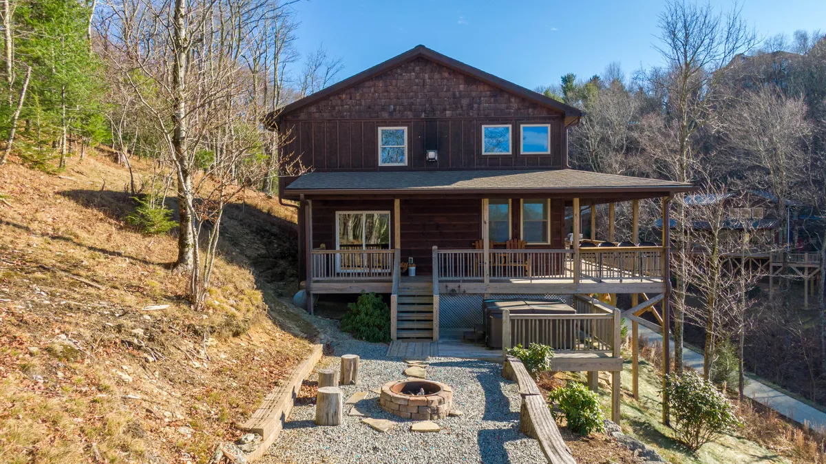 An inviting outdoor space with a wooden deck, built-in fire pit, and surrounding comfortable chairs, all framed by rock landscaping and vibrant trees. The scene is perfect for group gatherings and enjoying sunsets or starlit evenings.
