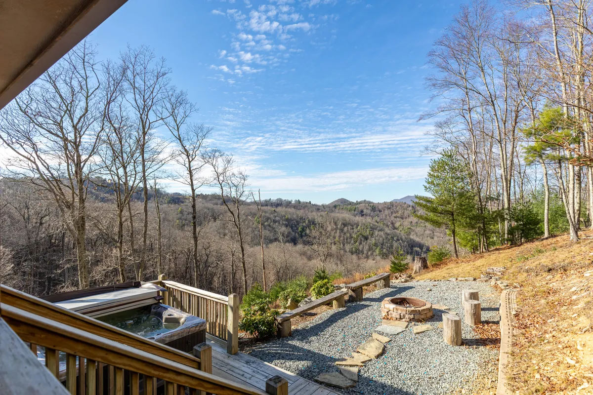 This image showcases the outdoor deck area featuring both the private hot tub and the fire pit, set against a beautiful mountain backdrop. The inviting wooden deck offers seating, and the view highlights a serene, resort-style atmosphere with trees and open sky.