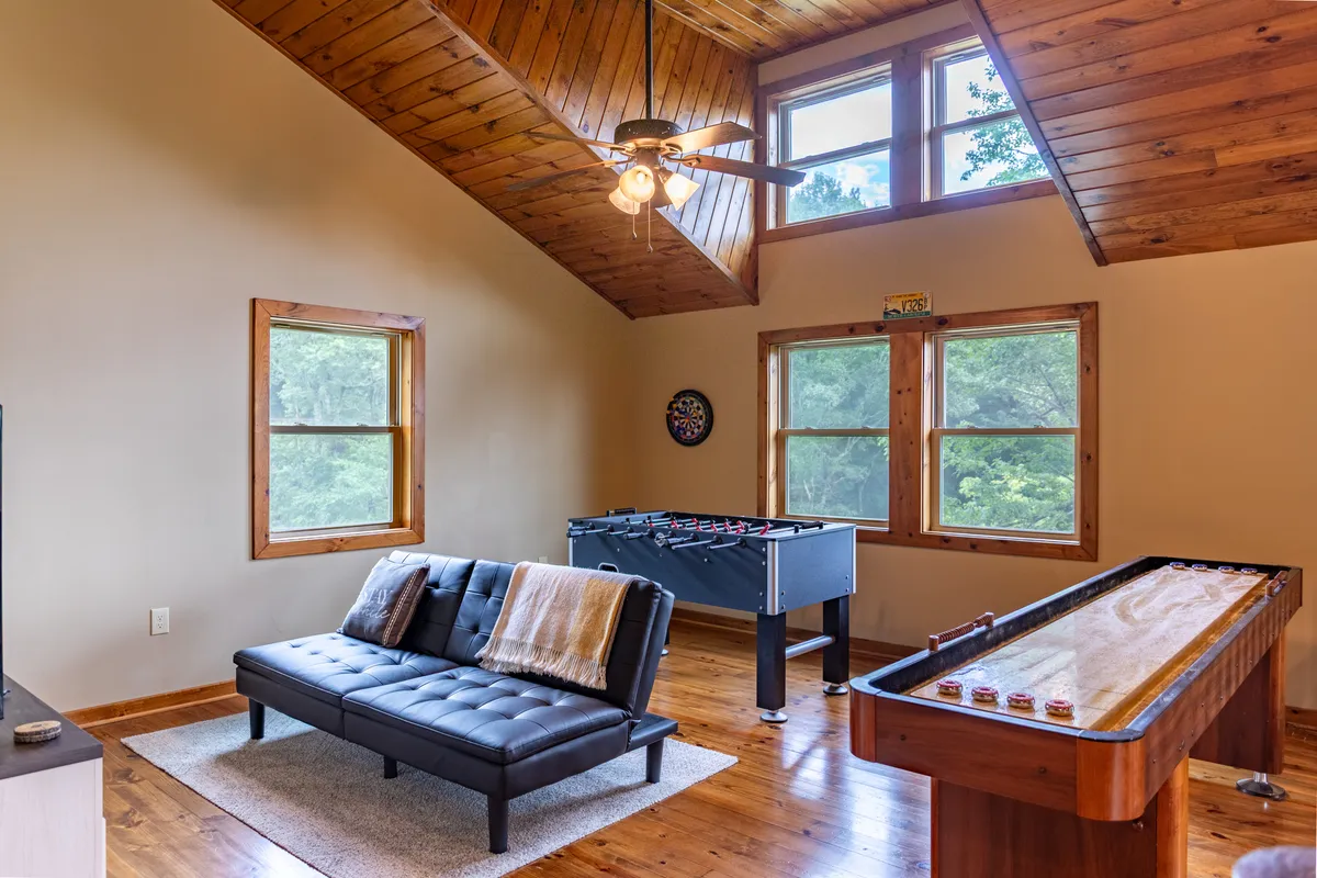 A vibrant game loft with a sofa for lounging, a full-size foosball table, a shuffleboard table, and windows letting in natural light. The space exudes a playful, casual vibe ideal for friendly competition and group fun.