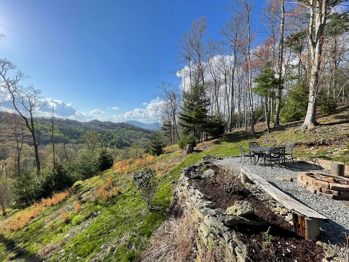 A tranquil outdoor seating area surrounds a cozy fire pit, set against distant mountain views and a tree-lined backdrop. The setup suggests both relaxation and evening gatherings, making it ideal for roasting marshmallows or sharing stories under the stars.