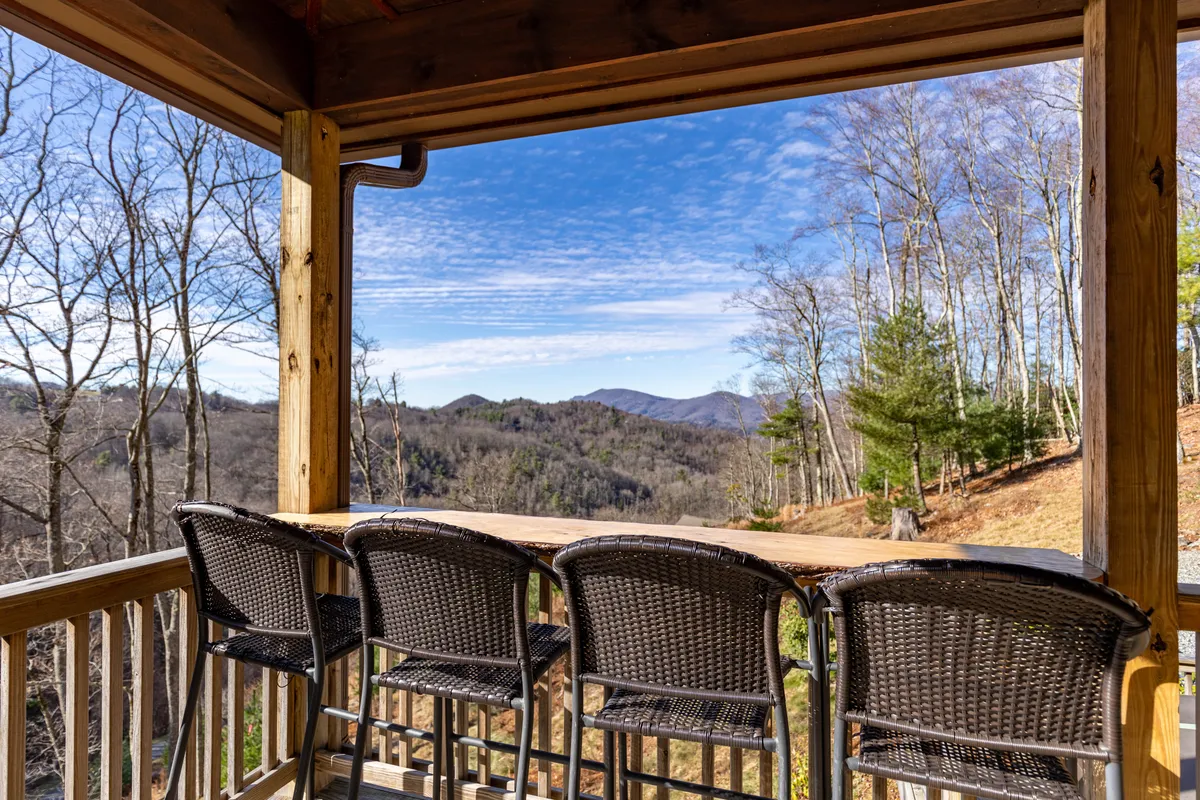 A tranquil scene featuring a live-edge bar with seating on the covered porch, overlooking a sweeping mountain view and tree line. The setup is ideal for morning coffee or evening drinks, with a seamless connection to nature.