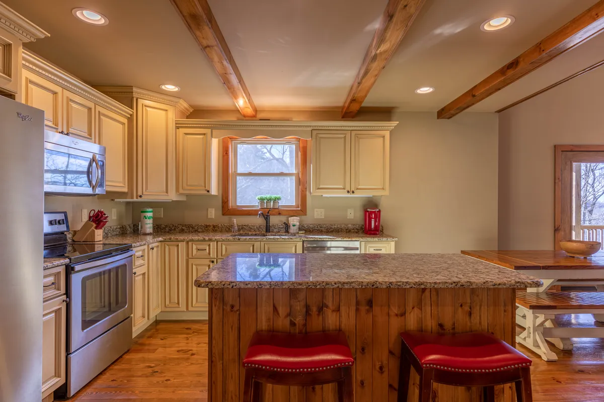 Featuring a generous kitchen island with granite countertops, red bar stools, and abundant natural light streaming in through a window, this image captures a cozy, lively atmosphere. The visible wooden beams and adjacent kitchen workspace reinforce the welcoming, communal aspect.