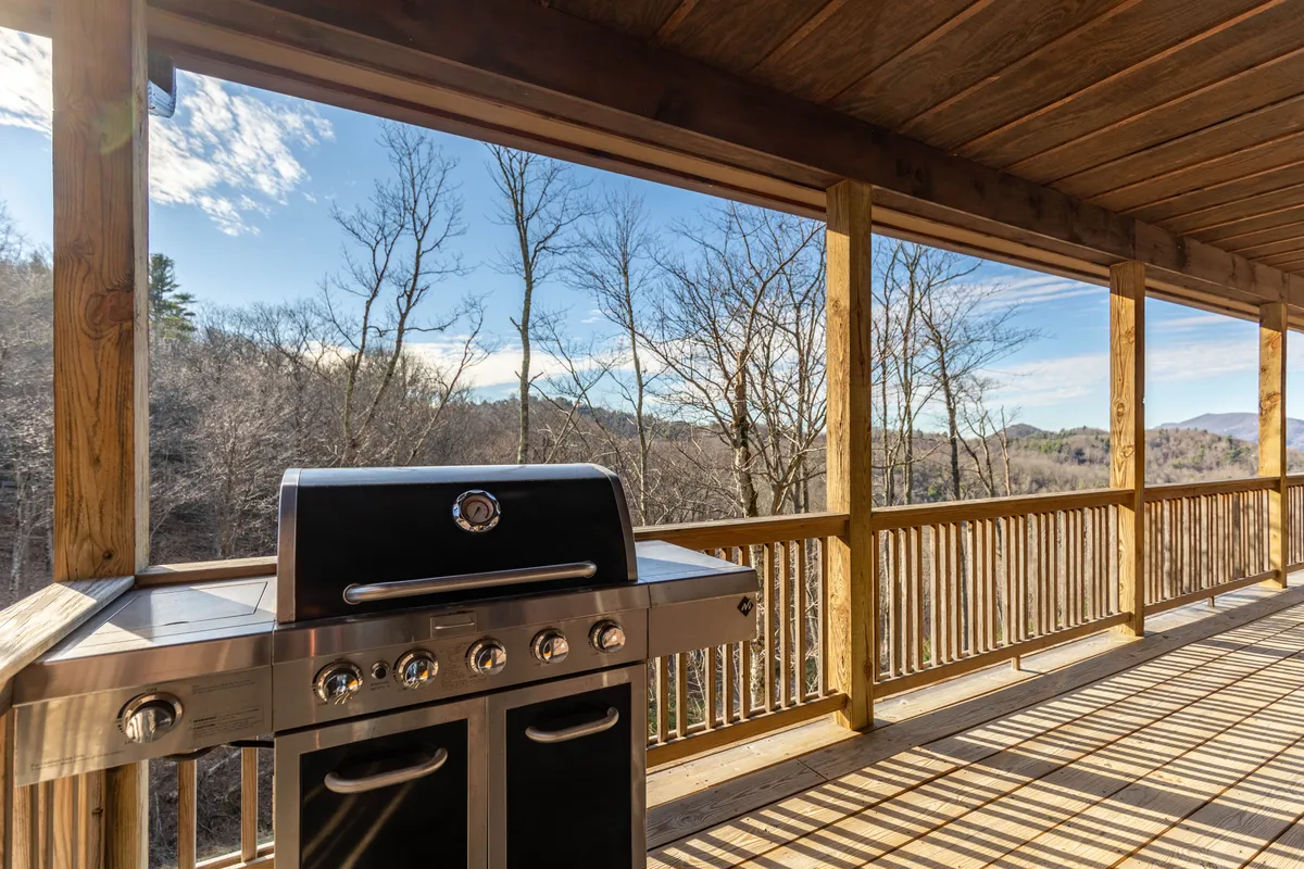 This exterior shot captures a deck with a grill and ample seating, set against a backdrop of lush trees and mountain scenery. The space is clearly designed for outdoor dining, barbecues, and group gatherings, with the mountains visible beyond the deck railing.
