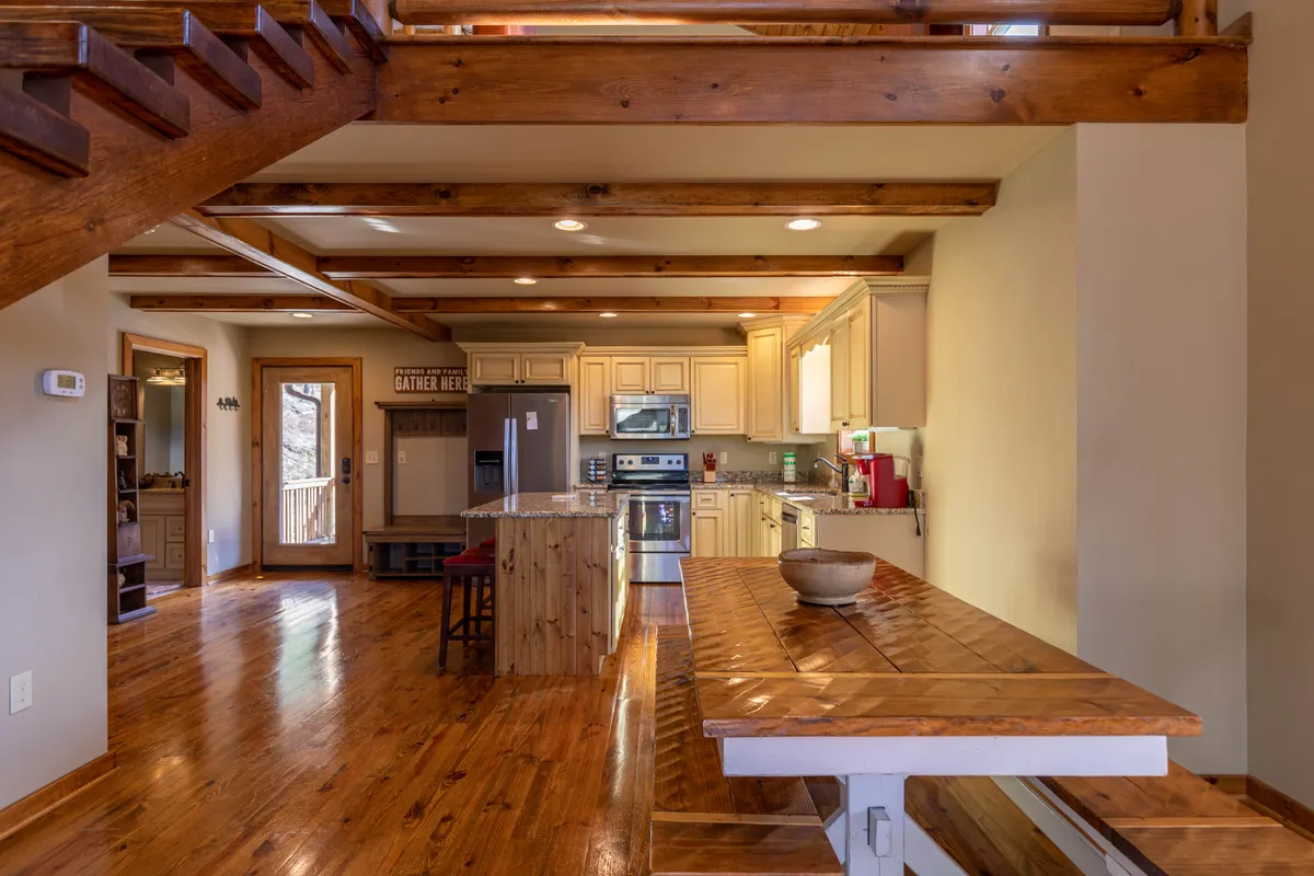 This interior scene highlights both the kitchen (with stainless steel appliances and a kitchen island) and the open dining area with a large wooden table. The open staircase in the background enhances the sense of space and flow between culinary and communal zones.