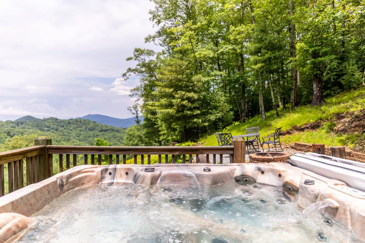 This image showcases a spacious outdoor deck featuring a private hot tub, surrounded by trees and offering panoramic mountain views. Comfortable seating is arranged nearby, inviting guests to relax and socialize while immersed in the natural beauty of the High Country.